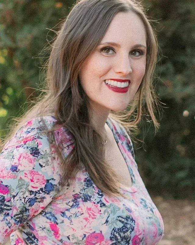 A young woman with long brown hair, smiling, wearing a floral top, outdoors with greenery in the background.