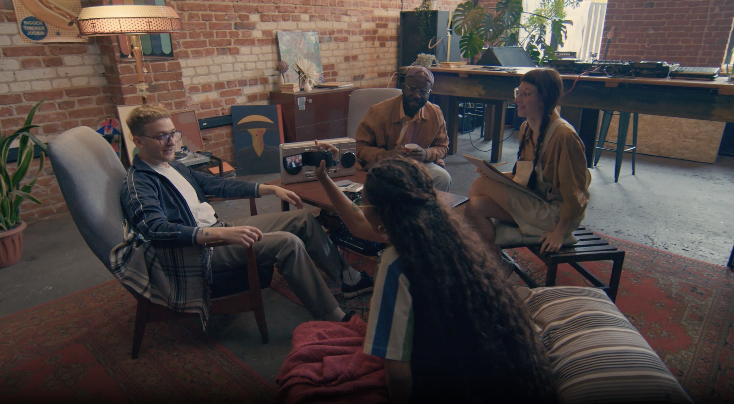 Group of five young adults having a casual discussion in a trendy loft with brick walls and eclectic decor, one person holding a smartphone and another with a clipboard.