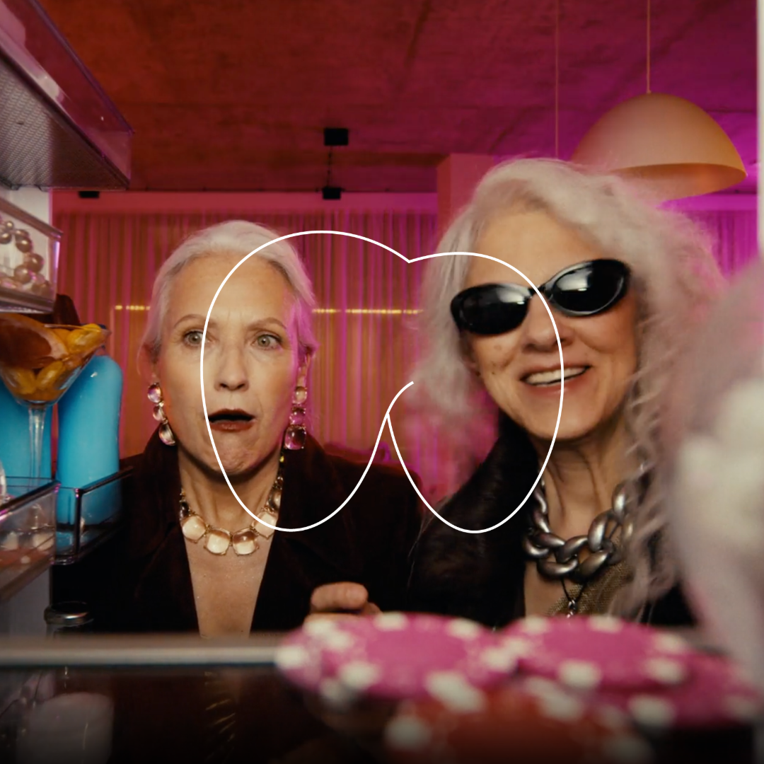 Two older women with platinum blonde hair at a casino, one with surprised expression and the other wearing sunglasses and smiling, surrounded by poker chips and colorful decor.