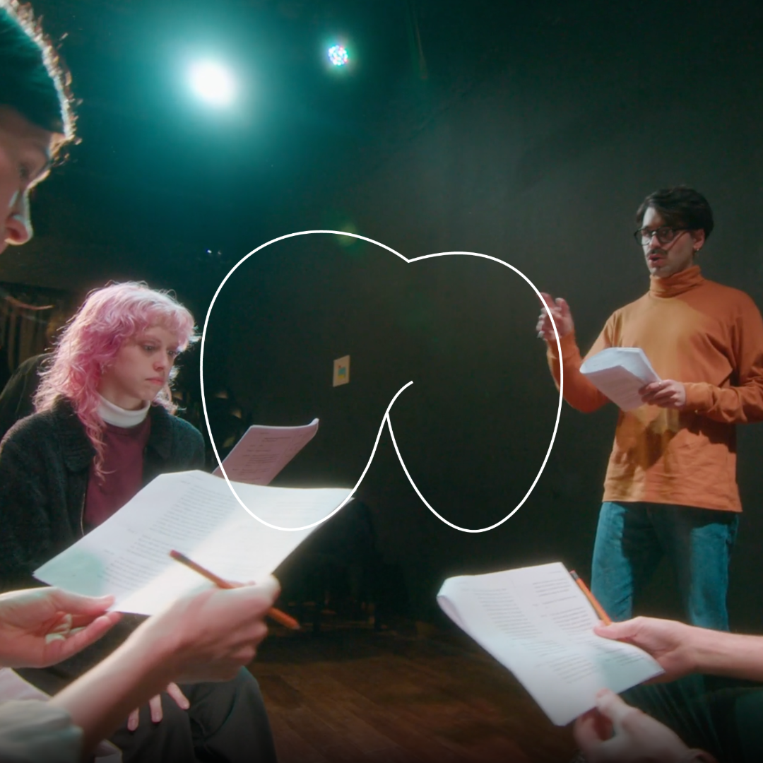 Group of people practicing script reading in a dark room with stage lighting, two women and one man holding papers, one woman with pink hair and the man with glasses and an orange turtleneck.