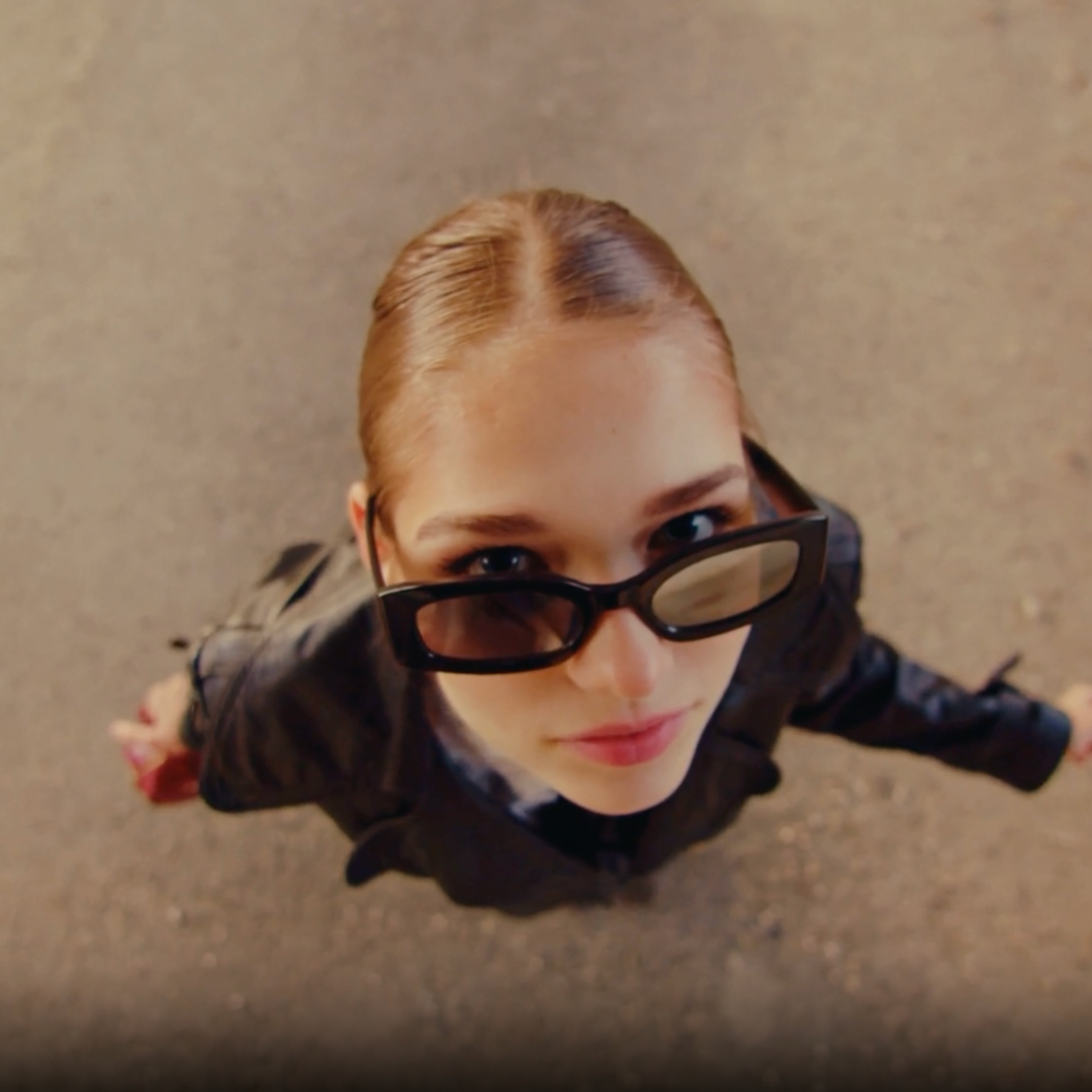 Overhead view of a woman with blond hair, wearing black sunglasses and a black leather jacket, looking up at the camera on a textured ground surface.