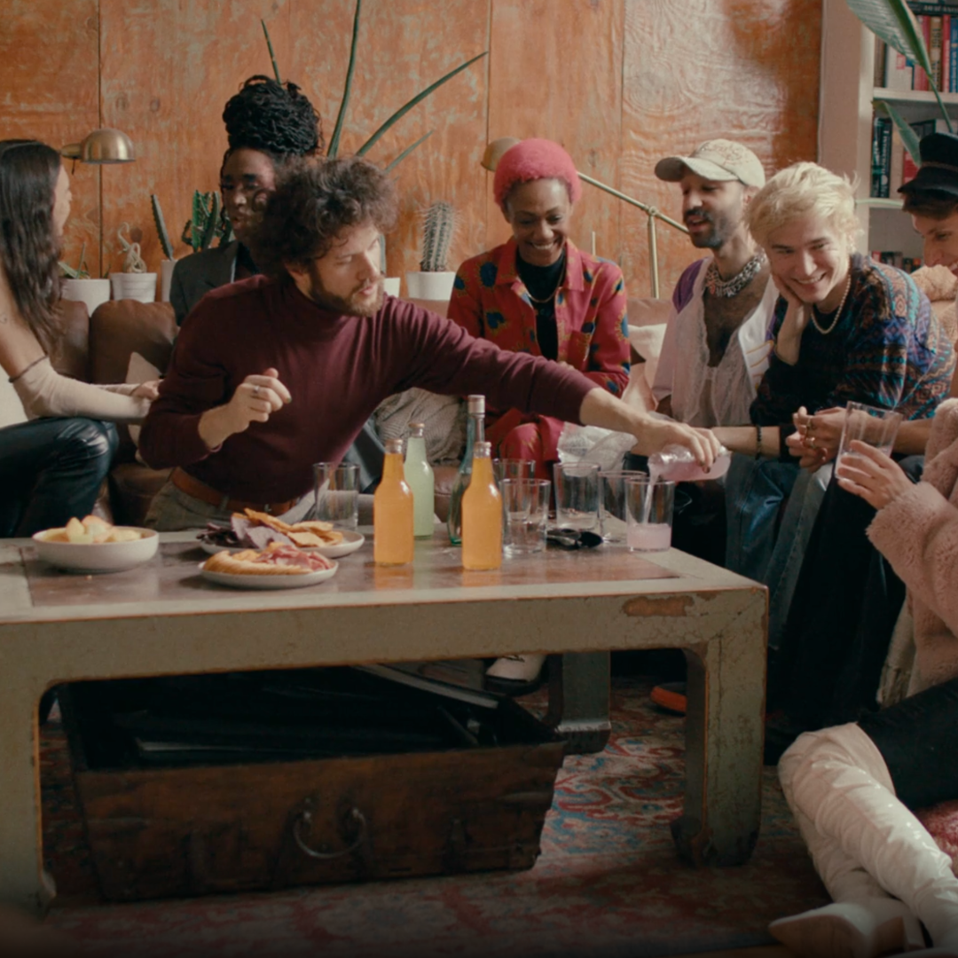 A group of diverse friends gathered around a coffee table enjoying drinks and snacks at a house party.