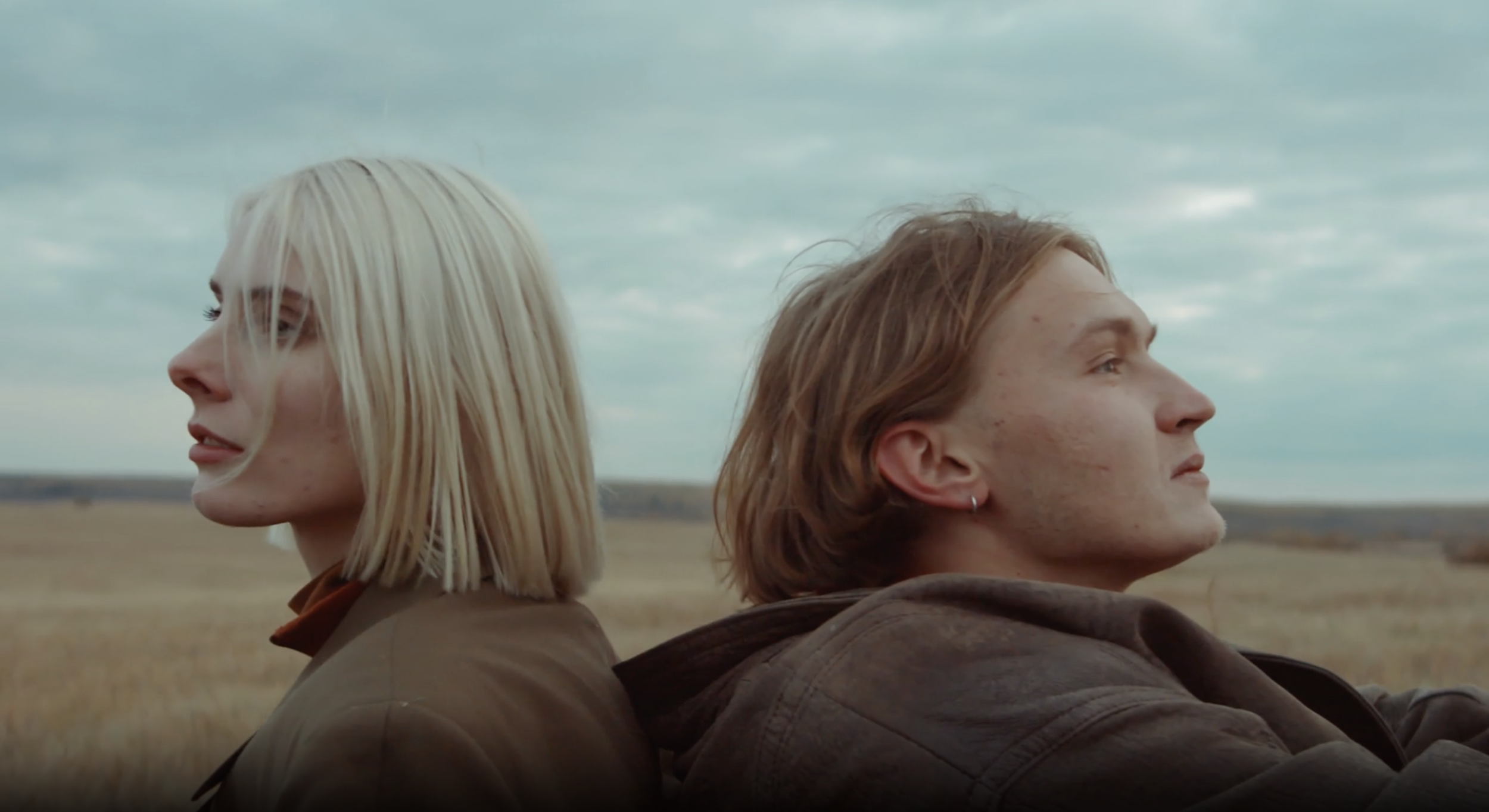 A man and woman sitting back-to-back outdoors in a field under a cloudy sky, facing opposite directions with calm expressions.
