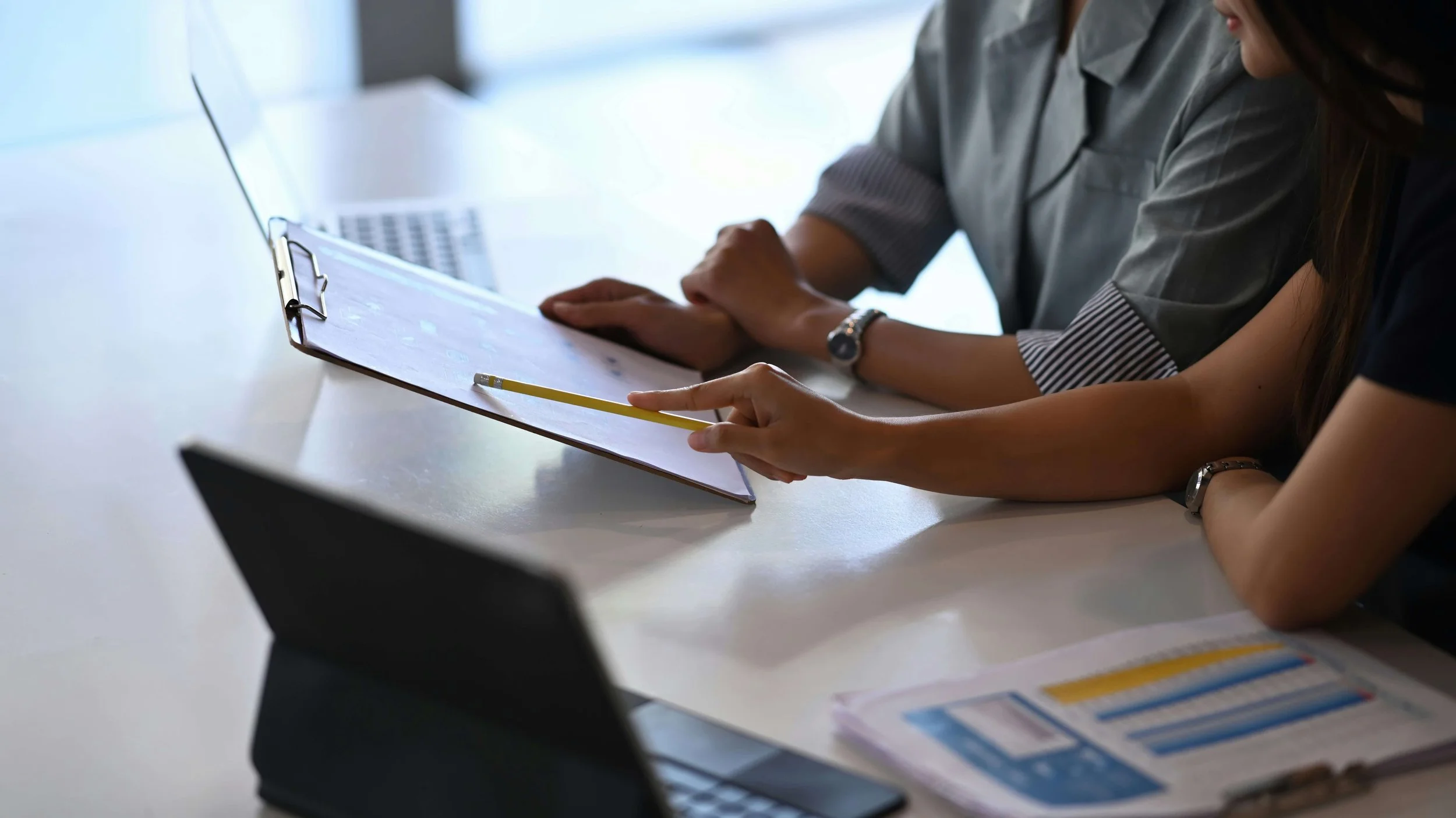 Two people reviewing documents on a clipboard at a desk with laptops and printed charts nearby.