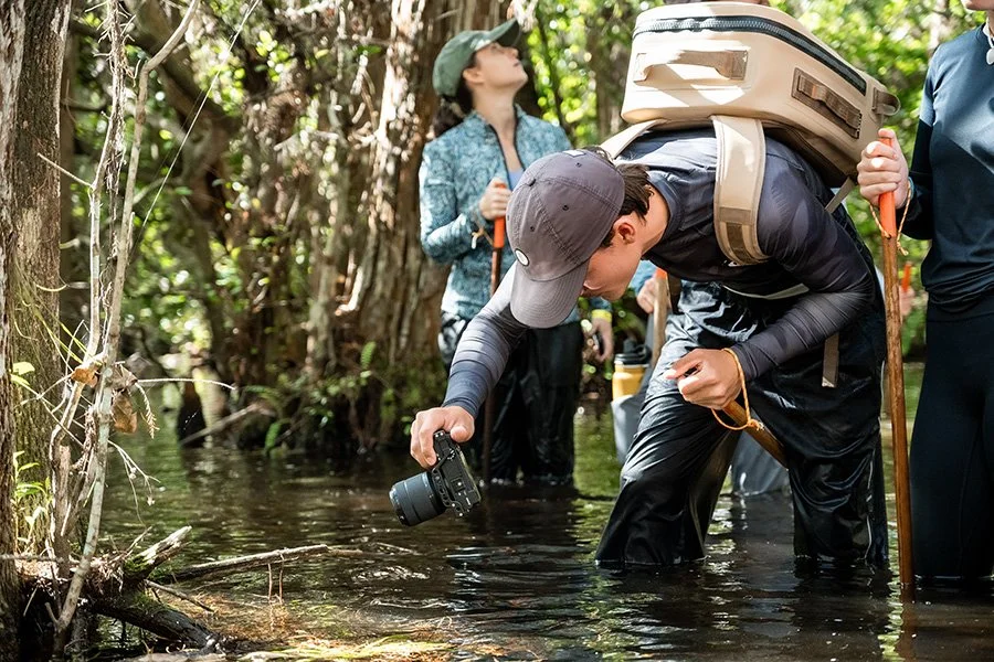 A group of people exploring a shallow stream in a forest, one person taking photos, others holding walking sticks, dressed in outdoor gear.