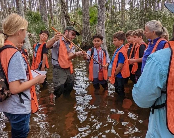 A group of children and two adults standing in a floodwater in a wooded area, with one adult demonstrating something to the children.