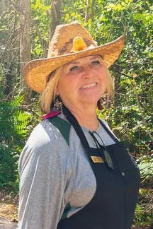 A woman wearing a wide-brimmed straw hat with a yellow flower, smiling outdoors in a wooded area, dressed in a gray long-sleeve shirt and black apron.