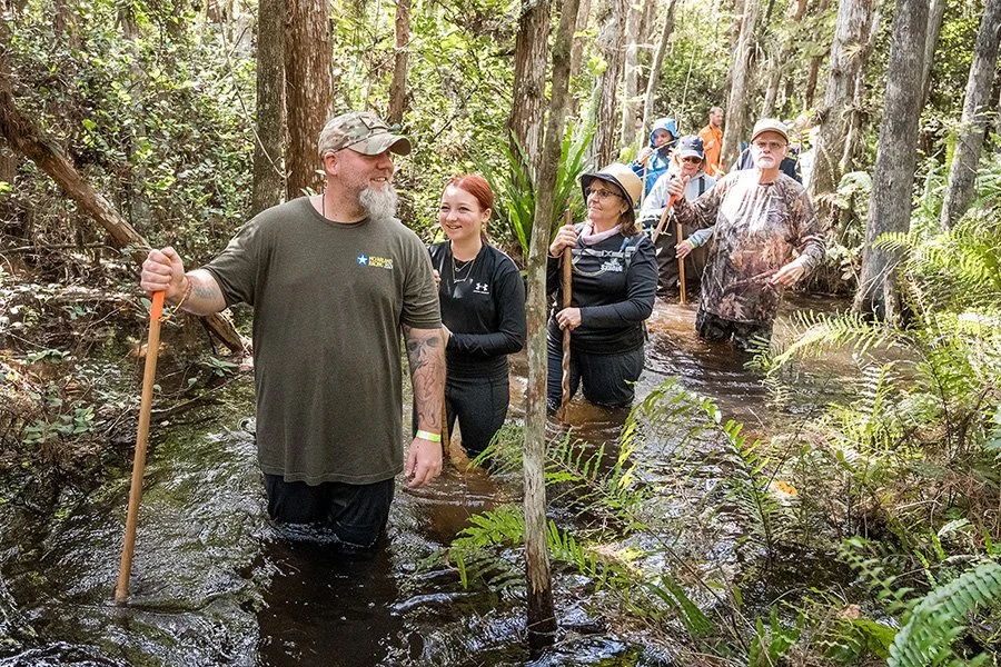 Group of people wading through a creek in a forest, some with walking sticks, enjoying a nature hike, smiling and chatting.