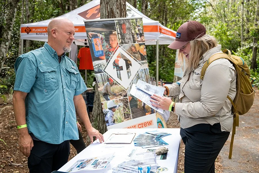 A woman with a backpack getting information from a man at an outdoor event booth about outdoor adventure activities, with a banner displaying images of people camping and hiking, and a tent in the background.