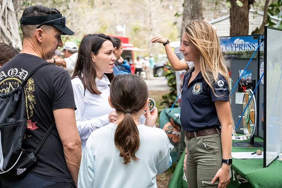 A group of people talking outdoors at an event, with a woman in a navy blue shirt explaining something to a man, woman, and girl, next to a booth with informational materials.