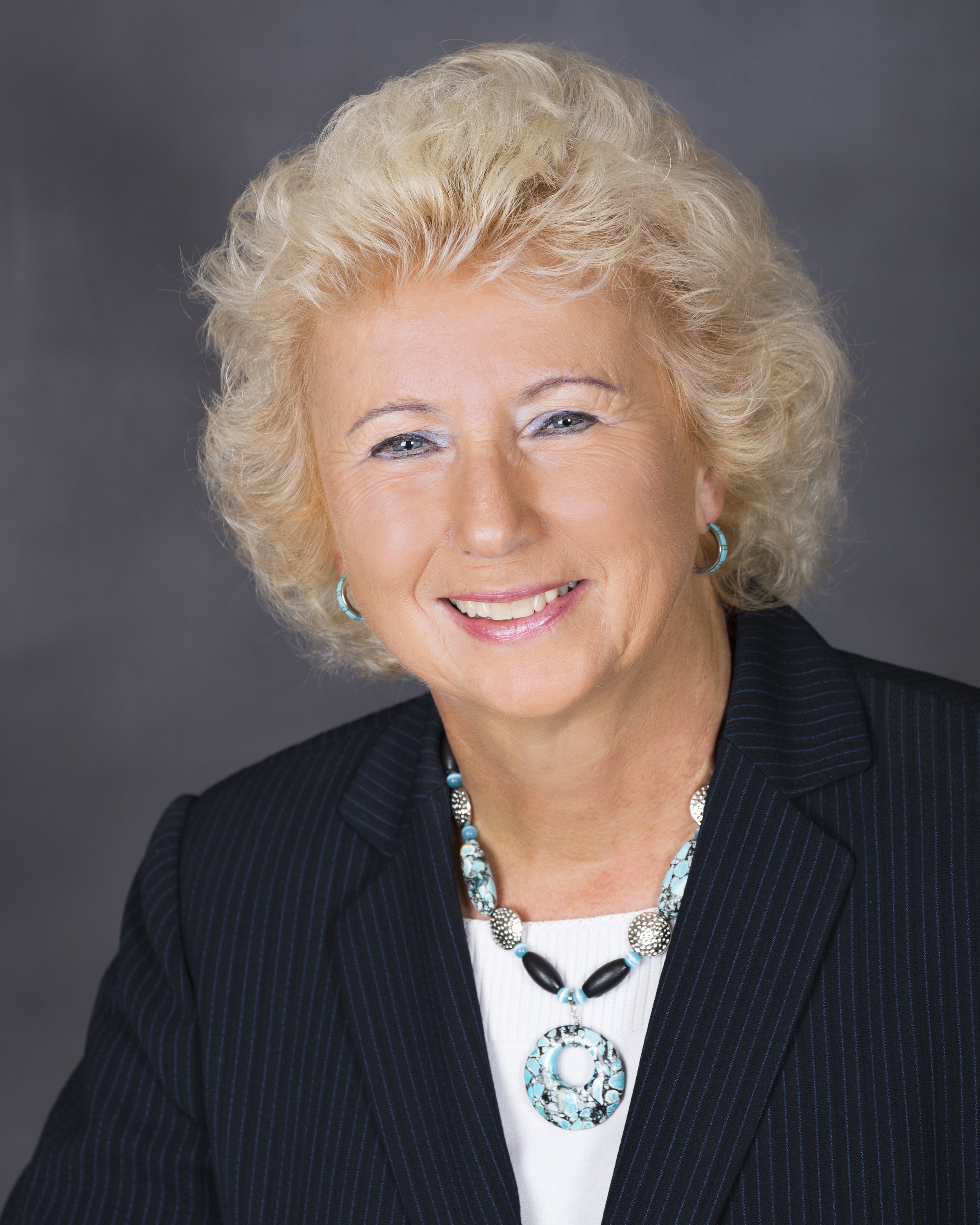 A smiling elderly woman with short, curly blonde hair, wearing a dark pinstripe blazer, white top, and turquoise jewelry, posed against a plain gray background.