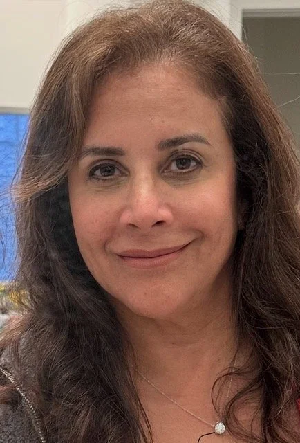A woman with light skin, long wavy brown hair, and a gentle smile, wearing a necklace with a small pendant, in an indoor setting.