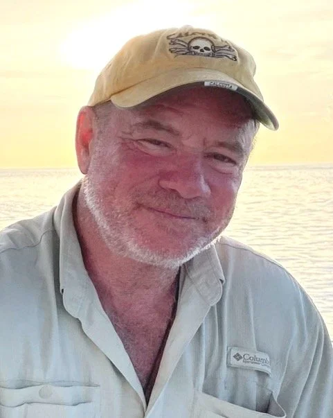 A man in a beige baseball cap with a skull design, smiling, with a sunset over water in the background.