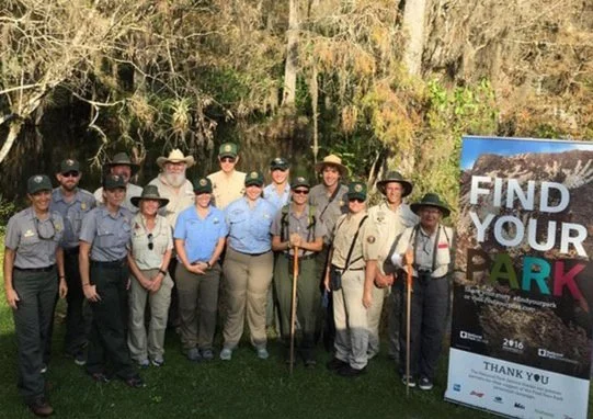 Group of 14 people, including park rangers, standing outdoors in front of trees and a sign that reads 'Find Your Park,' for a park event or celebration.