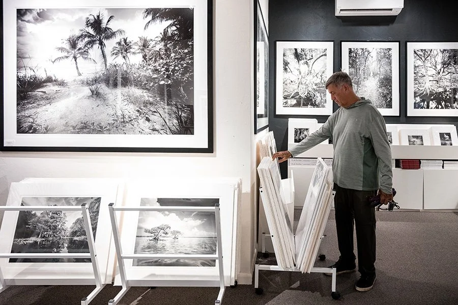 A man in a grey hoodie browsing framed black and white photographs in an art gallery, with more photos displayed on the walls and leaning on white racks.