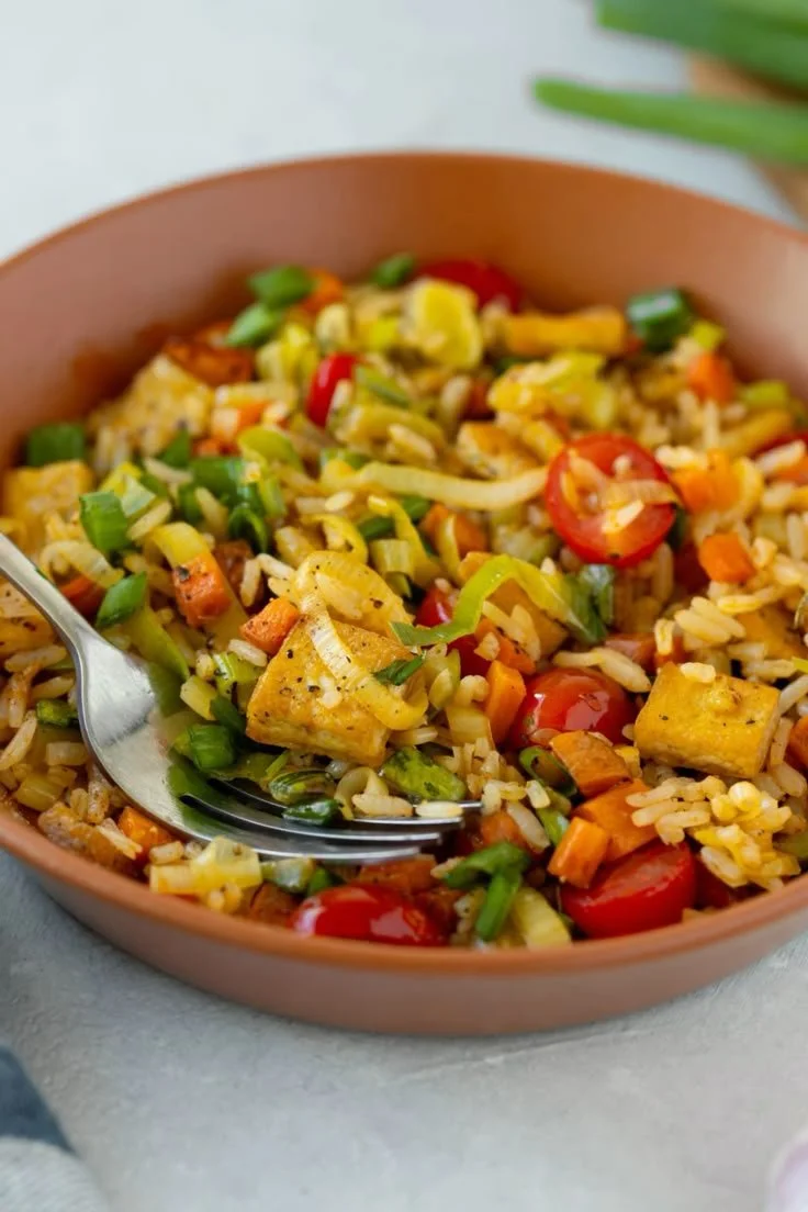 Piatto di riso fritto con tofu, verdure, pomodorini e cipollotti in una ciotola marrone.