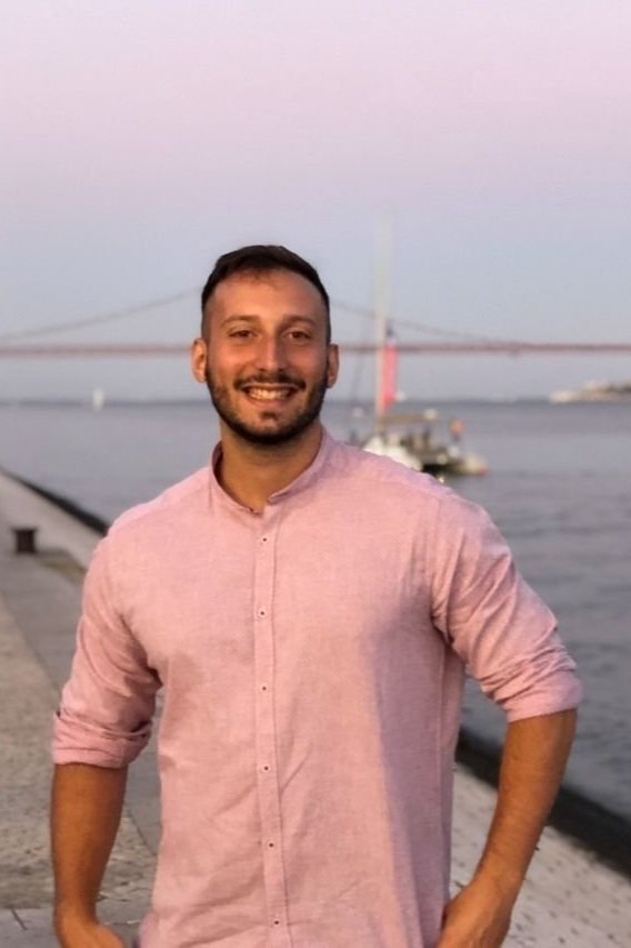 Persona sorridente con camicia rosa davanti a un ponte e un fiume al tramonto.