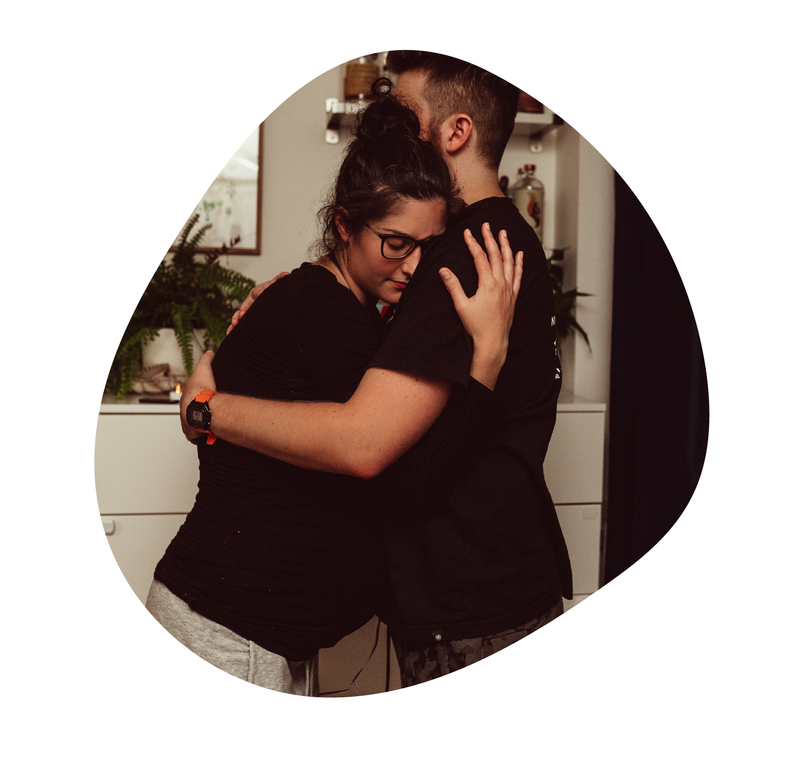 Two people hugging each other in a heartfelt embrace inside a room, with one person wearing glasses and a black shirt, the other wearing a black shirt and patterned leggings.