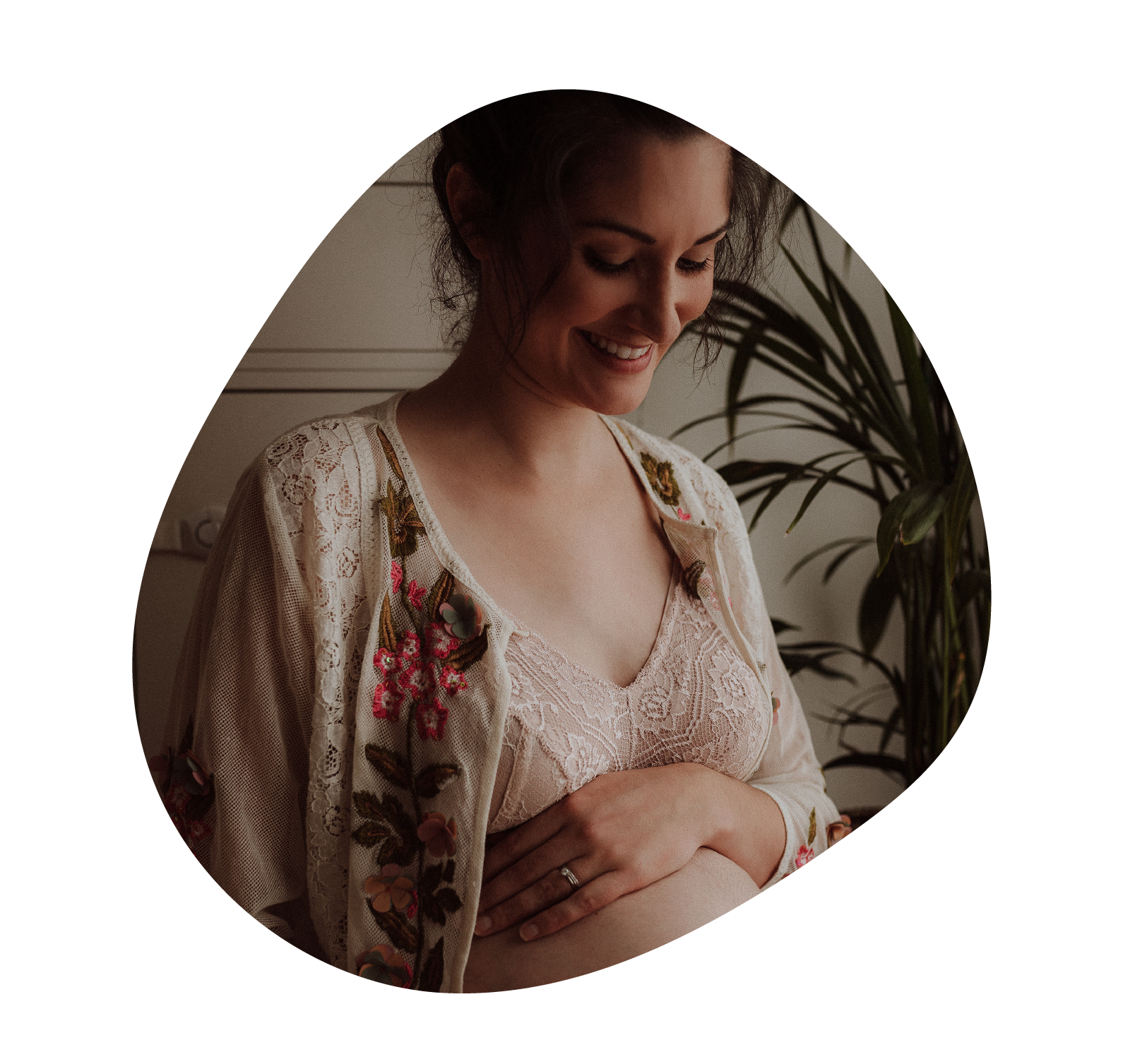 A smiling pregnant woman with light skin and dark hair, wearing a lace top and a floral embroidered kimono, gently holding her belly with both hands, indoors near a large houseplant.