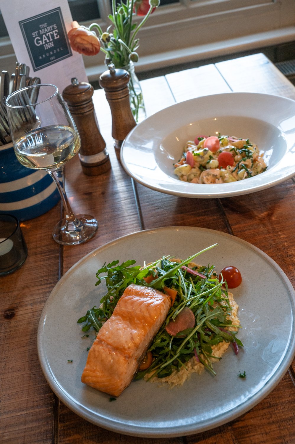 A table setting with a dish of grilled salmon on a bed of greens and couscous, and a bowl of pasta with vegetables. There is a glass of white wine, salt and pepper shakers, and a small vase of flowers.