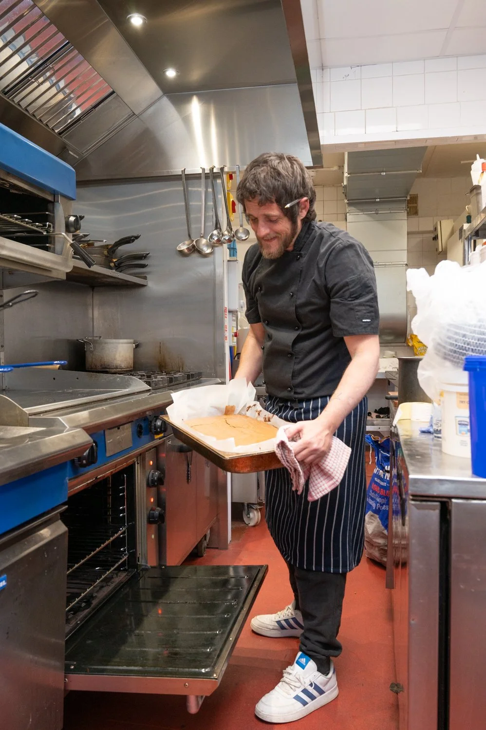 Chef placing a baking tray into an oven in a professional kitchen, wearing striped apron and sneakers.