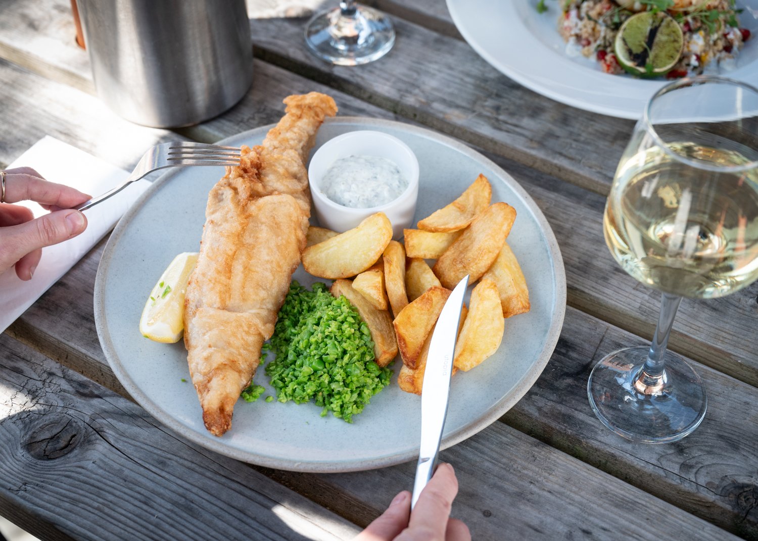 Plate of battered fish, thick potato wedges, mushy peas, tartar sauce, lemon wedge, with a glass of white wine on a wooden table.