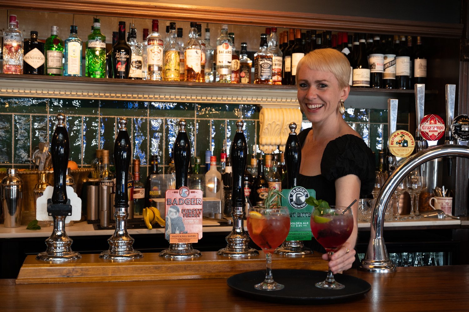 Smiling bartender in front of a bar with various liquor bottles, beer taps, and two cocktails on a tray.