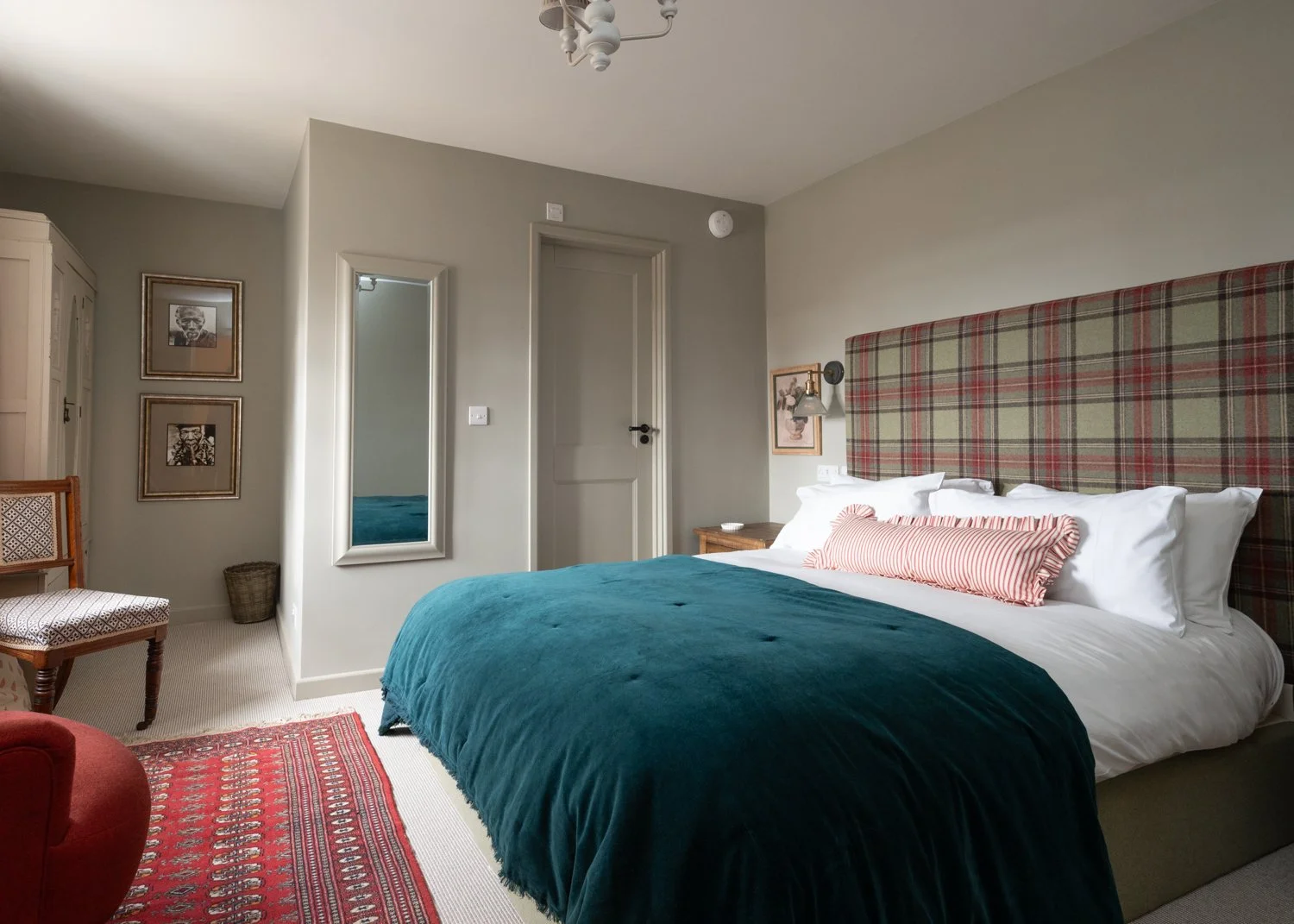 A cozy bedroom with a large bed featuring a plaid headboard and teal blanket. There's a striped pillow on white sheets. A red patterned rug covers part of the floor. The room includes a chair, a mirror, framed artwork on the wall, and a soft, neutral color scheme.