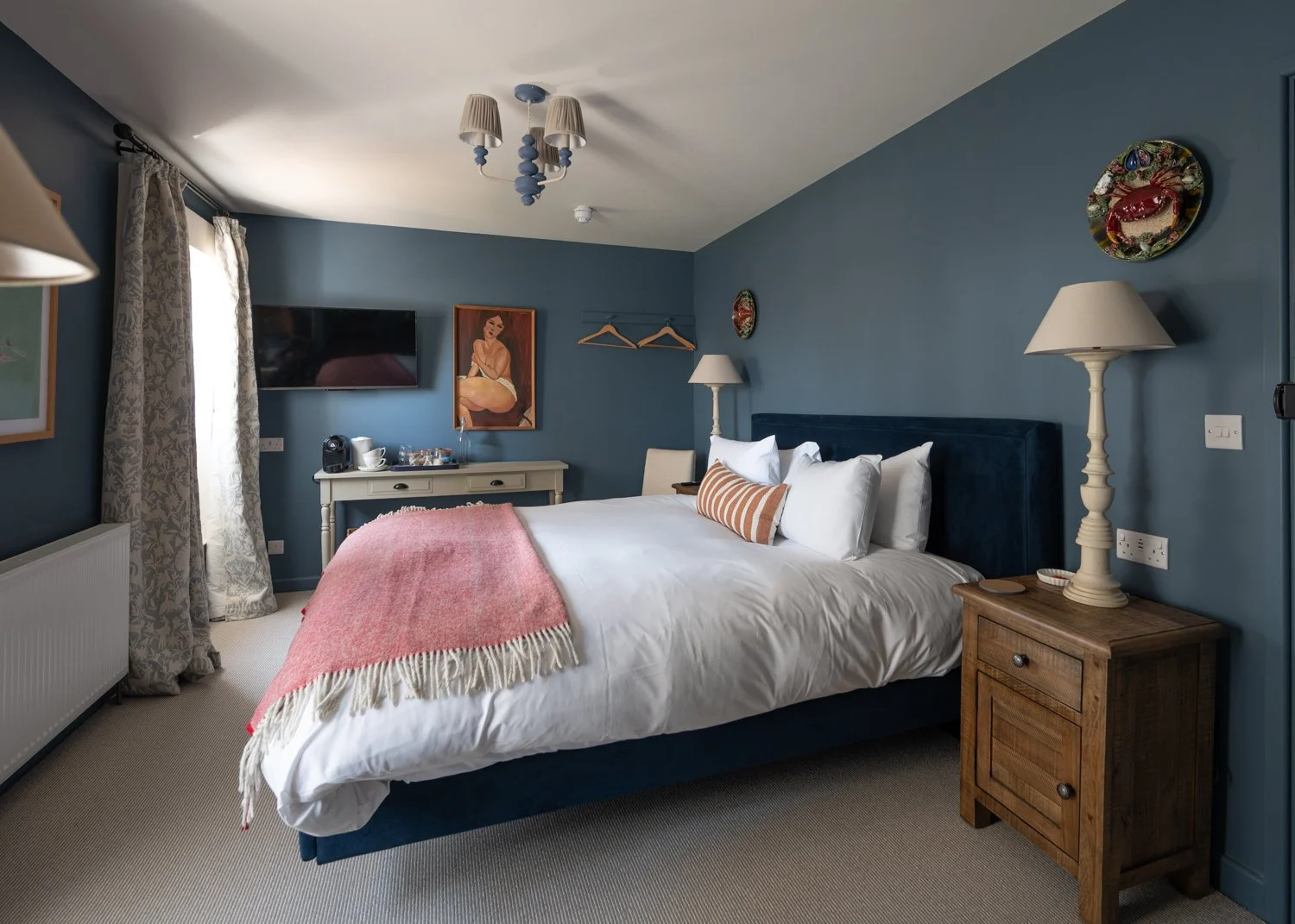 Cozy bedroom with blue walls, a white bed with a pink throw, wooden nightstand, desk with a coffee maker, wall-mounted TV, and decorative artwork.