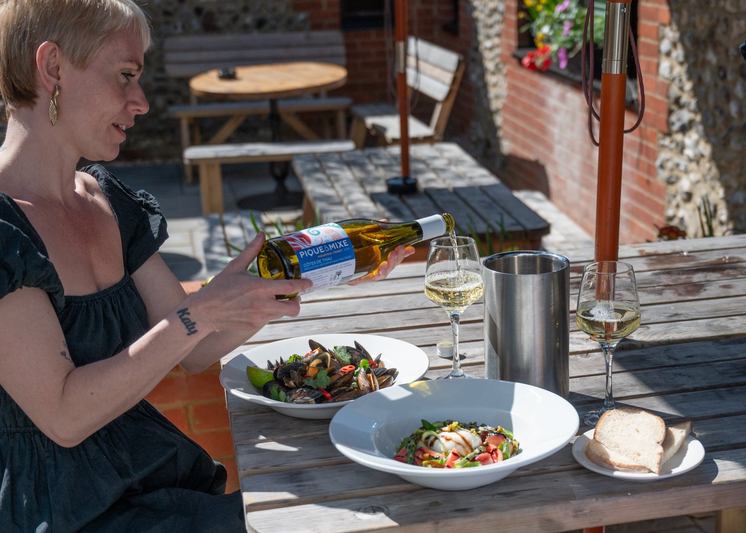 Person pouring white wine outdoors on a wooden table with dishes of seafood, pasta, and sliced bread in a garden setting.