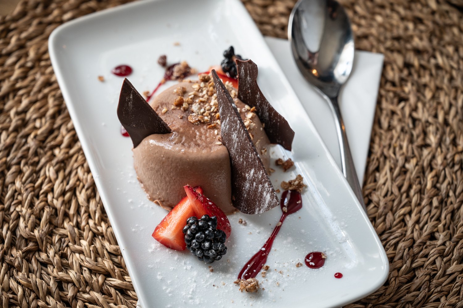 Chocolate panna cotta dessert with chocolate shards, strawberries, blackberries, nuts, and berry sauce on a white plate.