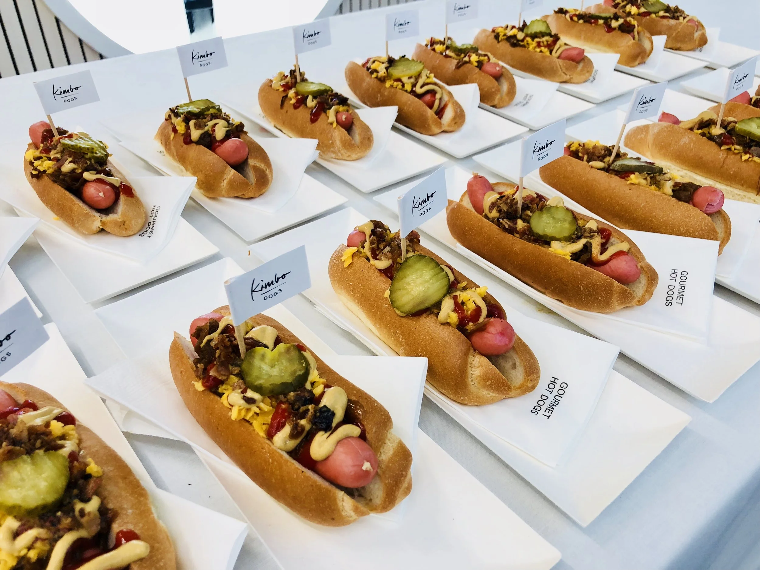 Mehrere Hotdogs auf Tellern, garniert mit Gurkenscheiben, Senf und weiteren Belägen, mit kleinen Fähnchen versehen.