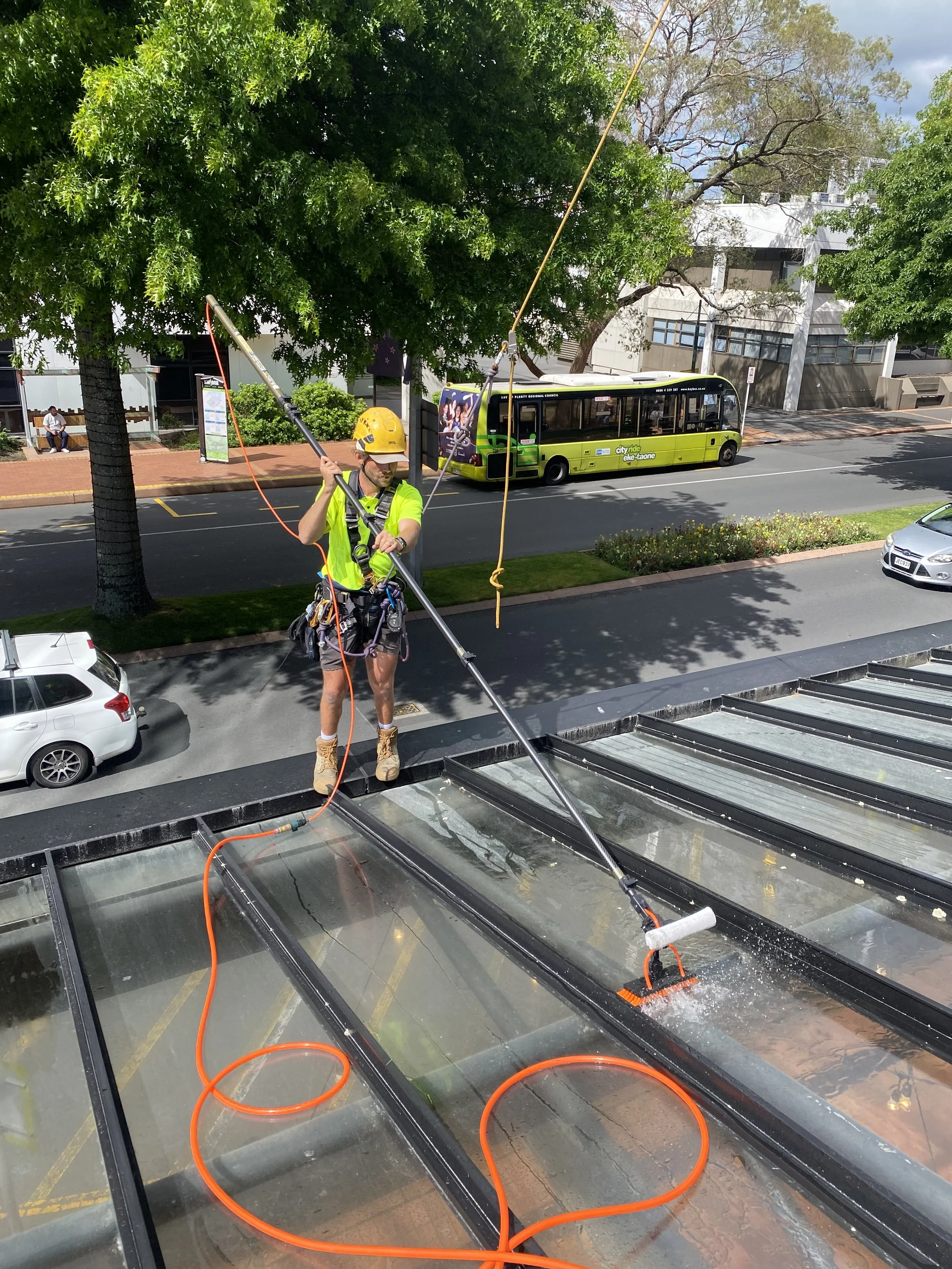 Worker wearing a yellow helmet and safety gear using a long tool to clean or maintain a large glass roof or skylight on a building, with trees and a city bus in the background.