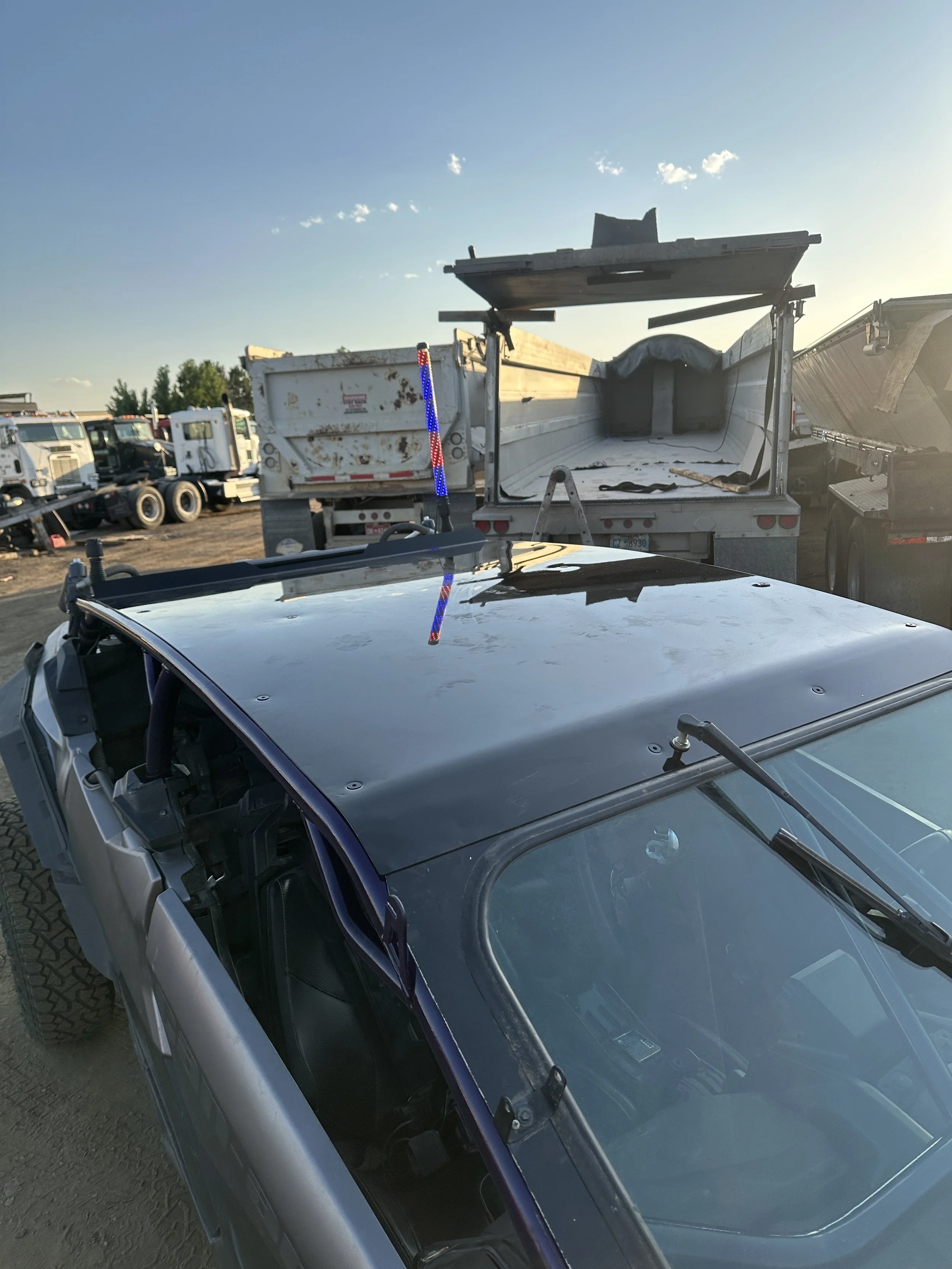 A car with a damaged roof parked in a lot with multiple trucks, including a dump truck and a flatbed truck, under a clear sky.