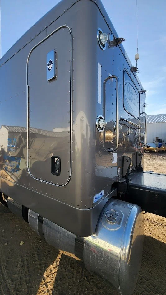 Close-up of a shiny silver semi-truck trailer with multiple lights and a door, parked outdoors on dirt ground under a clear blue sky.