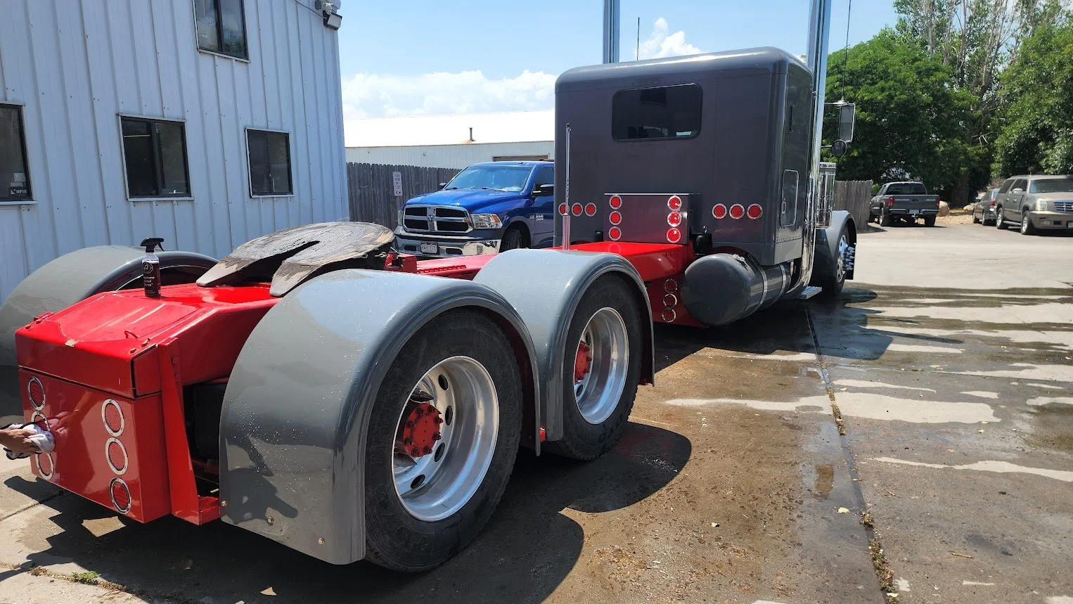 A semi-truck tractor being washed outside on a concrete surface, with water and soap visible on the ground. The tractor has a red and gray color scheme and is parked near a white building with multiple windows, with other vehicles in the background.