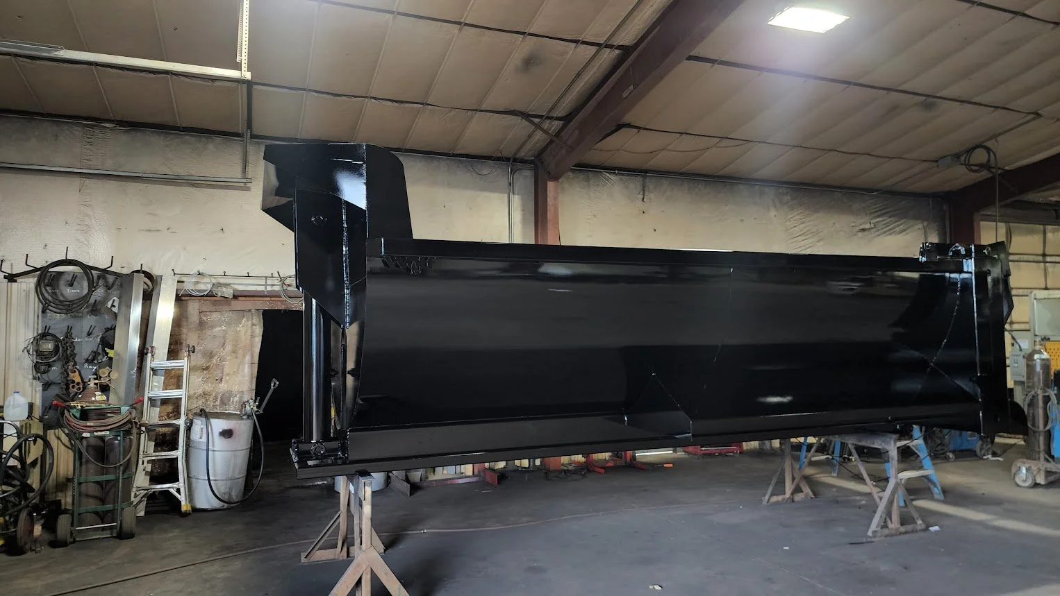 A large black metal structure in a workshop or factory setting, supported by wooden stands. The workshop has a high ceiling, fluorescent lights, and various tools and equipment lining the walls.