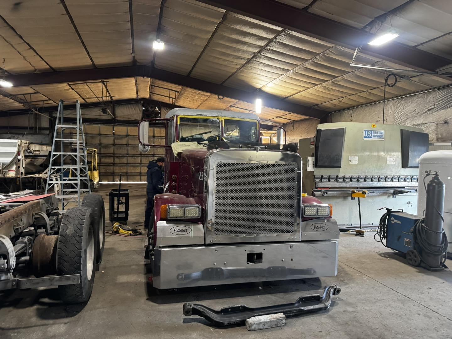 An indoor workshop with a vintage red semi-truck, another truck chassis on the left, and industrial equipment including a large U.S. industrial machinery machine, a welding machine, and a ladder.