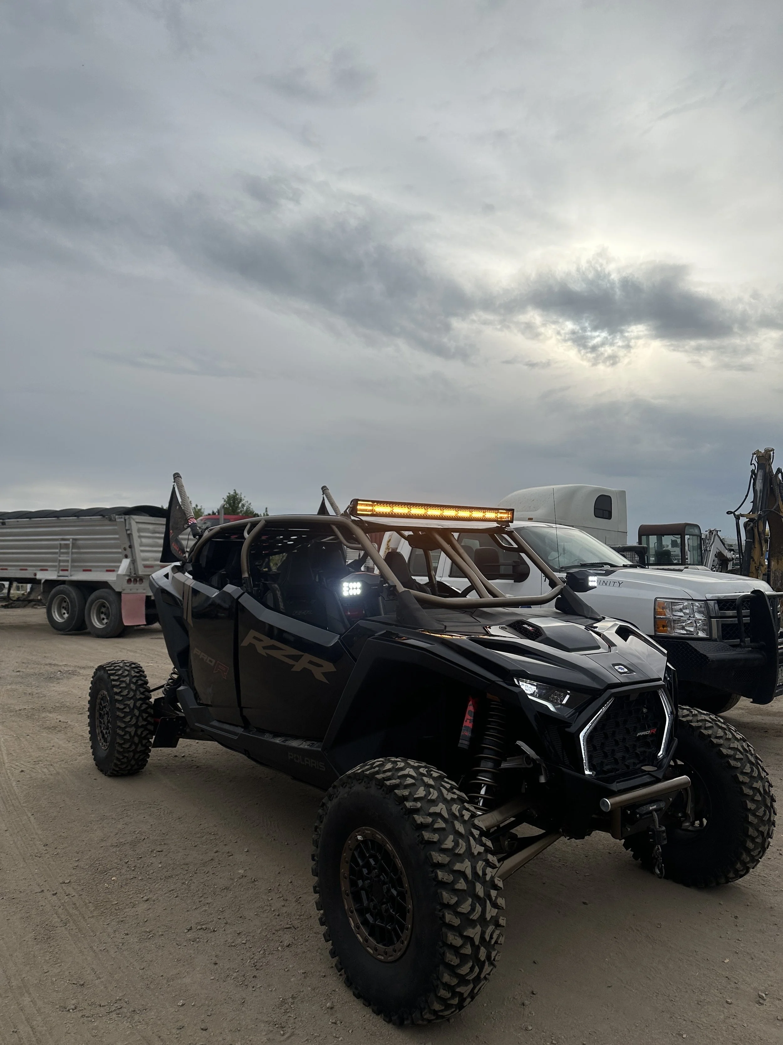 Black off-road Polaris RZR vehicle with large tires, roll cage, LED light bar, and driver side LED lights, parked on dirt with trucks and construction equipment nearby under a cloudy sky.