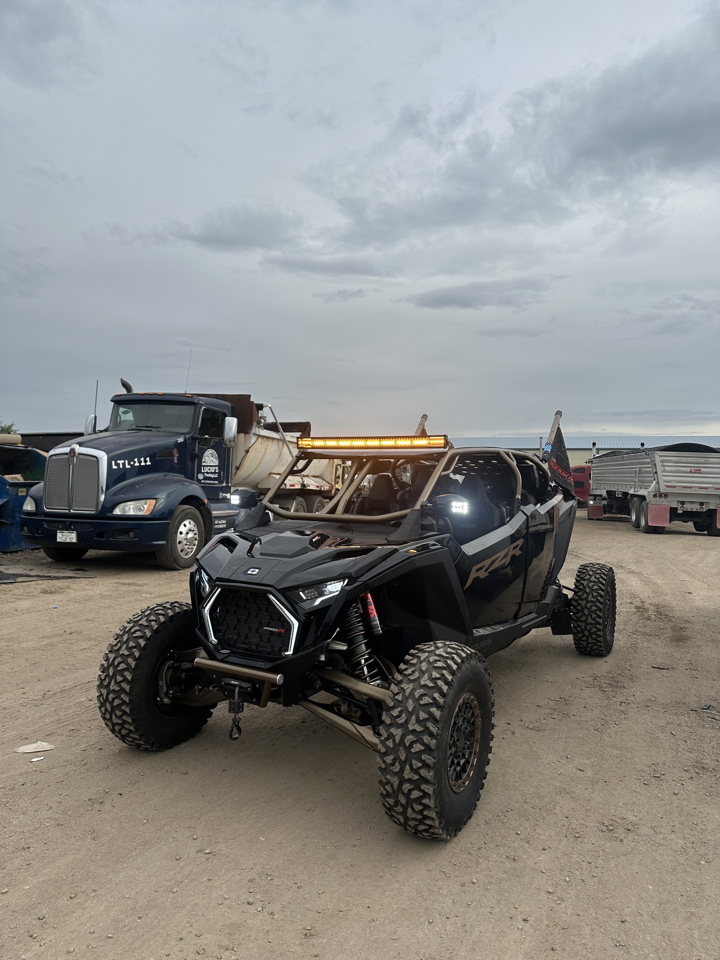 Black off-road RZR vehicle with large tires, a roof-mounted light bar, and a flag, parked on dirt ground with trucks in the background under a cloudy sky.