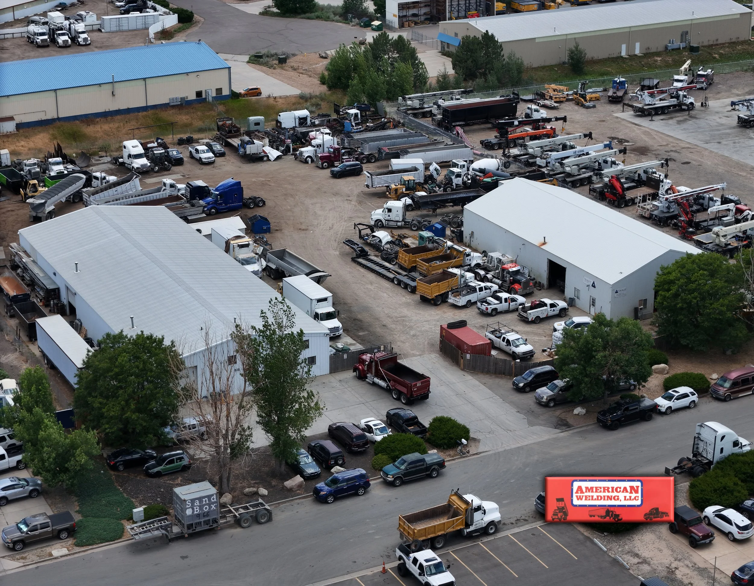 Aerial view of a commercial lot with various trucks, construction equipment, and trailers parked near metal buildings, with cars in a parking lot and trees in the foreground. Billboard reads 'American Welding, LLC'.