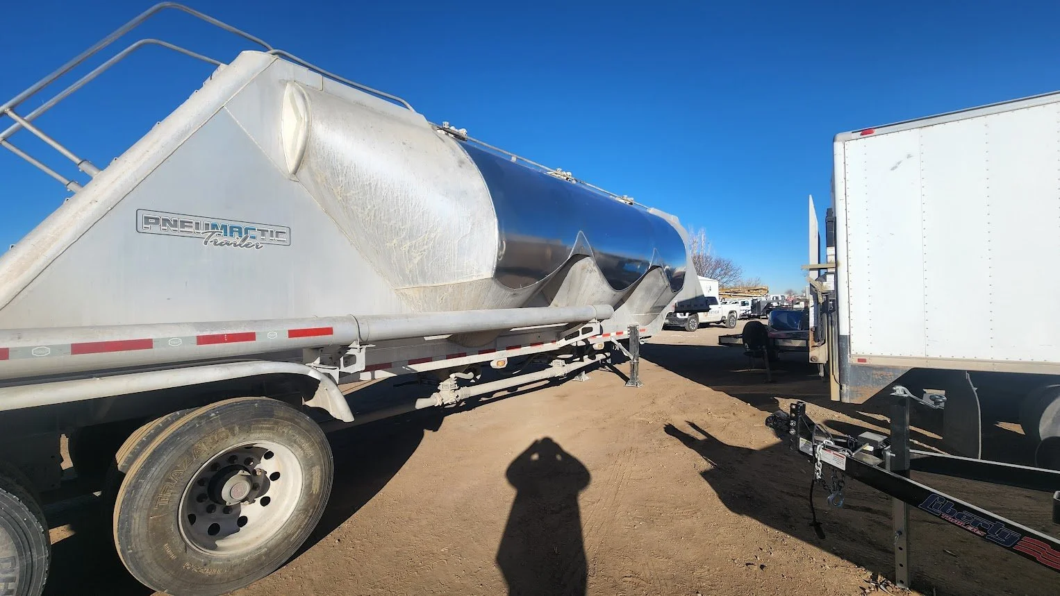 A large silver pneumatic trailer parked on a dirt lot, with several trucks and vehicles in the background under a clear blue sky.