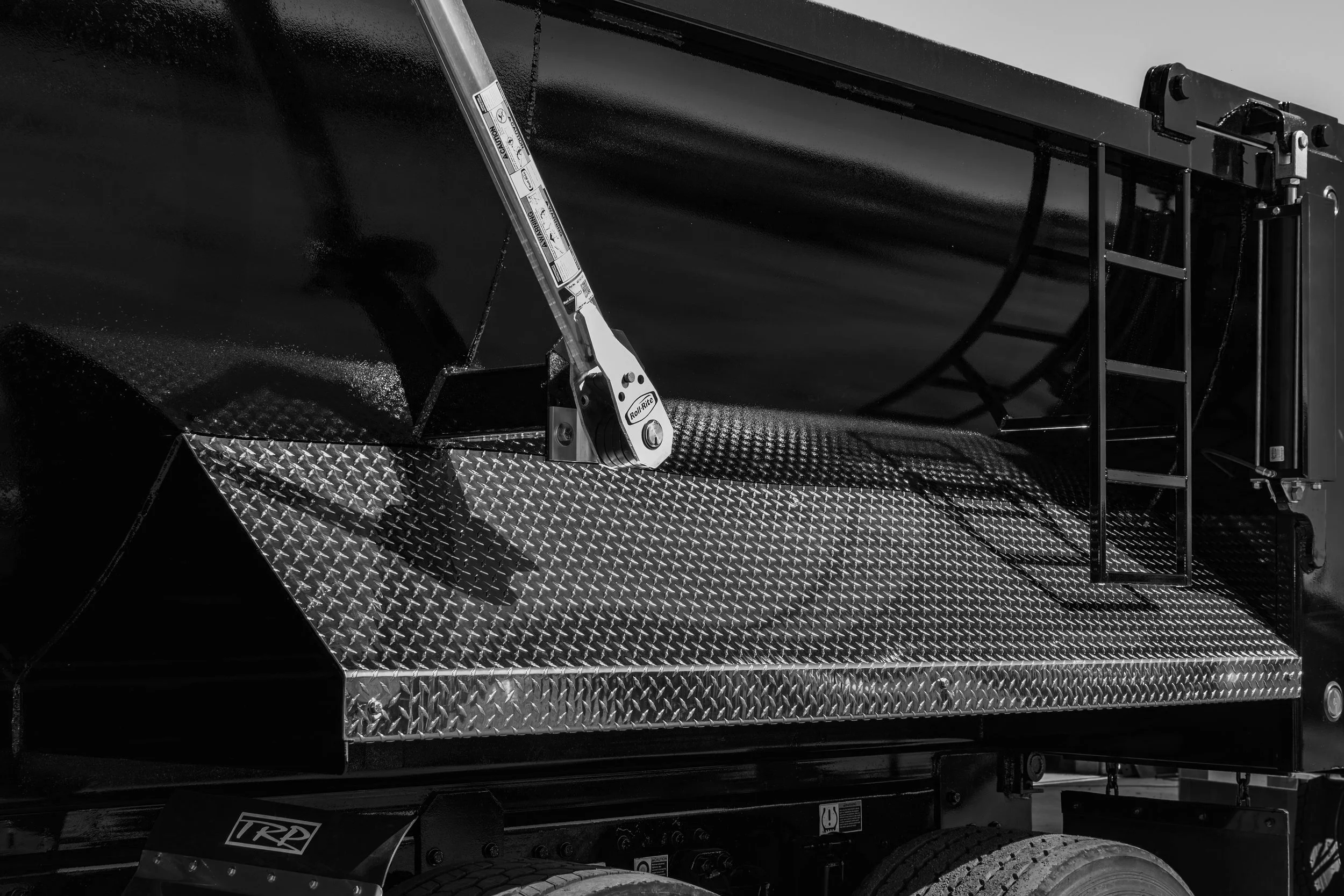 Close-up of a metal ramp or platform with textured diamond plate surface, part of a vehicle with a ladder, and a ratchet strap.