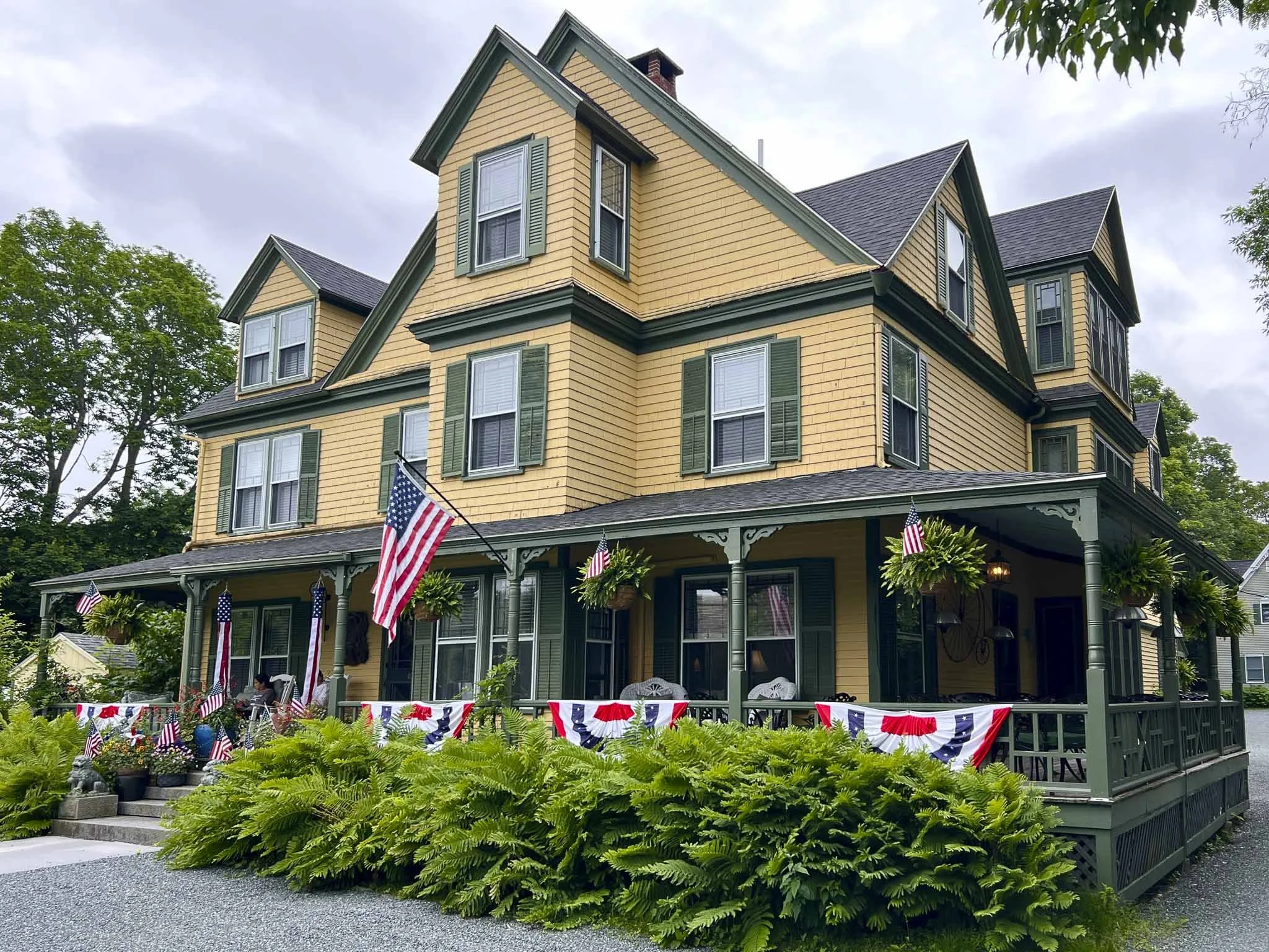 Victorian-style yellow house with green trim, American flag, wraparound porch, and hanging ferns