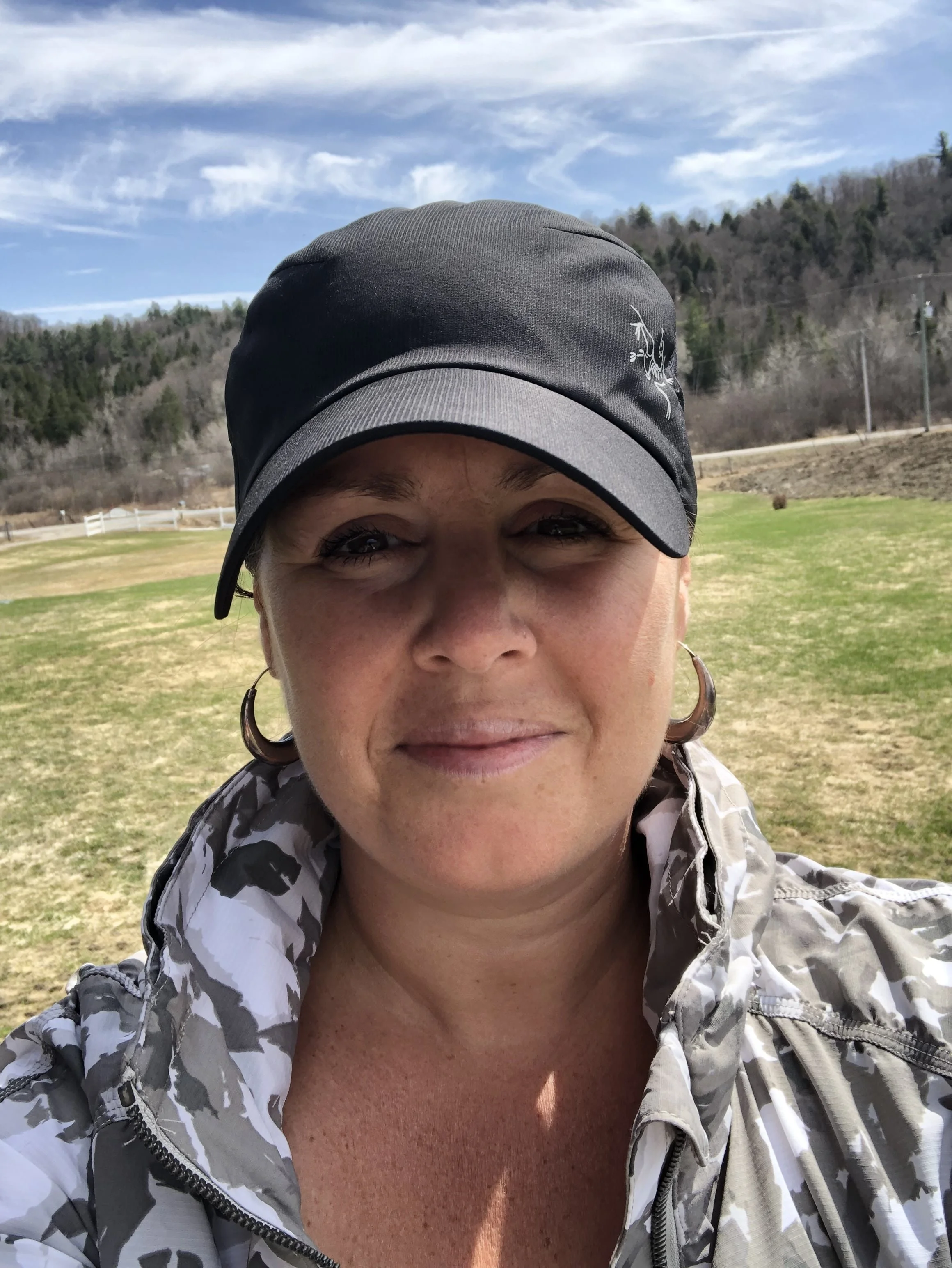 Woman in camouflage jacket and black cap standing outdoors with a grassy field and tree-covered hills in the background.