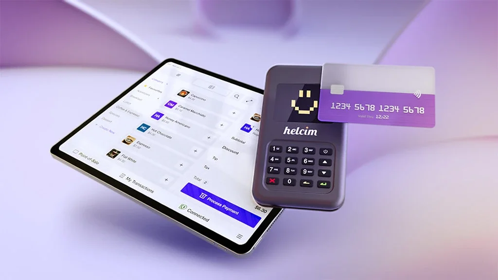 A tablet displaying the Helcim point-of-sale software interface next to a Helcim physical card reader and a credit card.