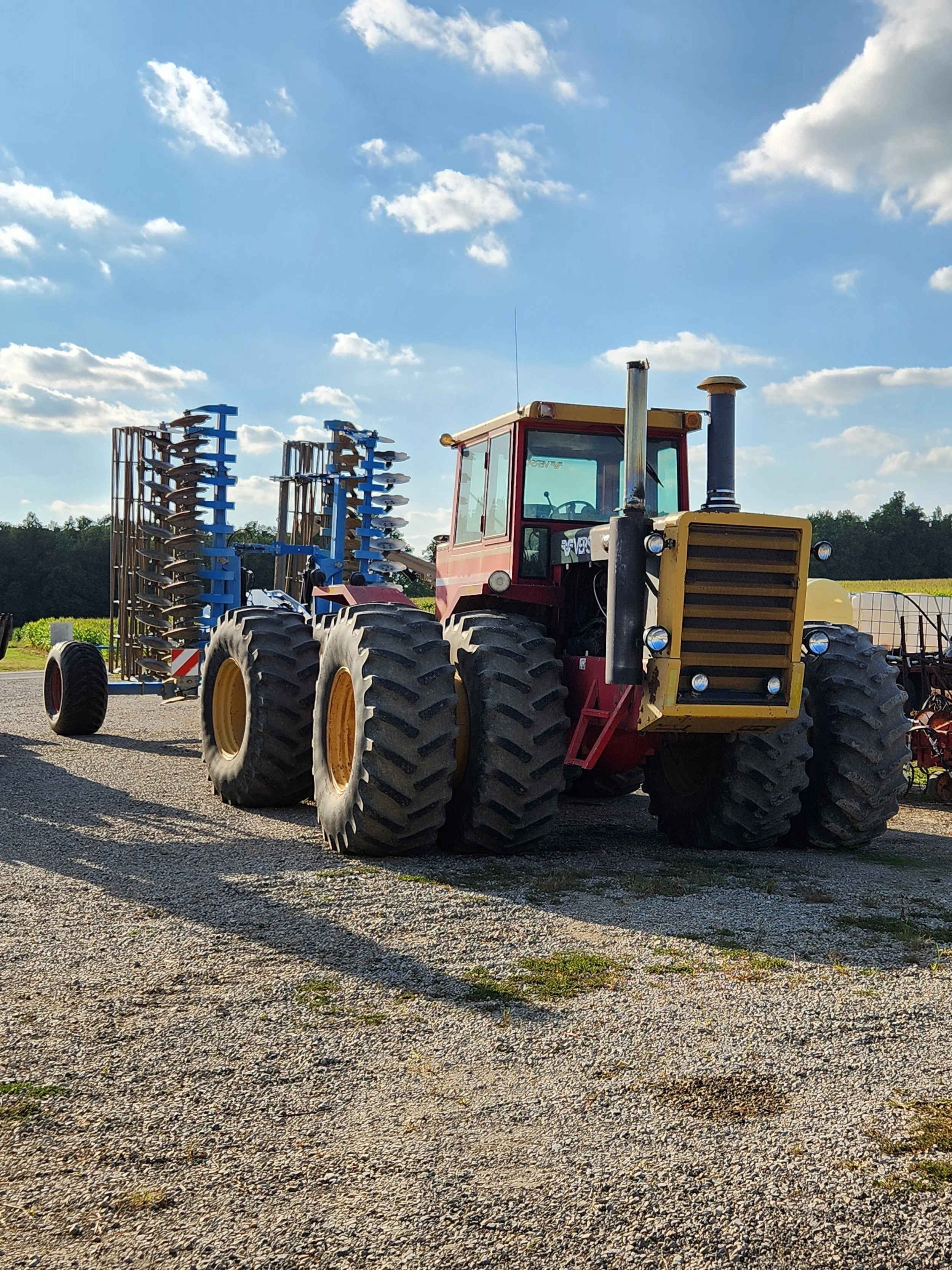 A large red and yellow tractor with multiple tires attached to agricultural equipment, parked on a gravel surface under a partly cloudy sky.