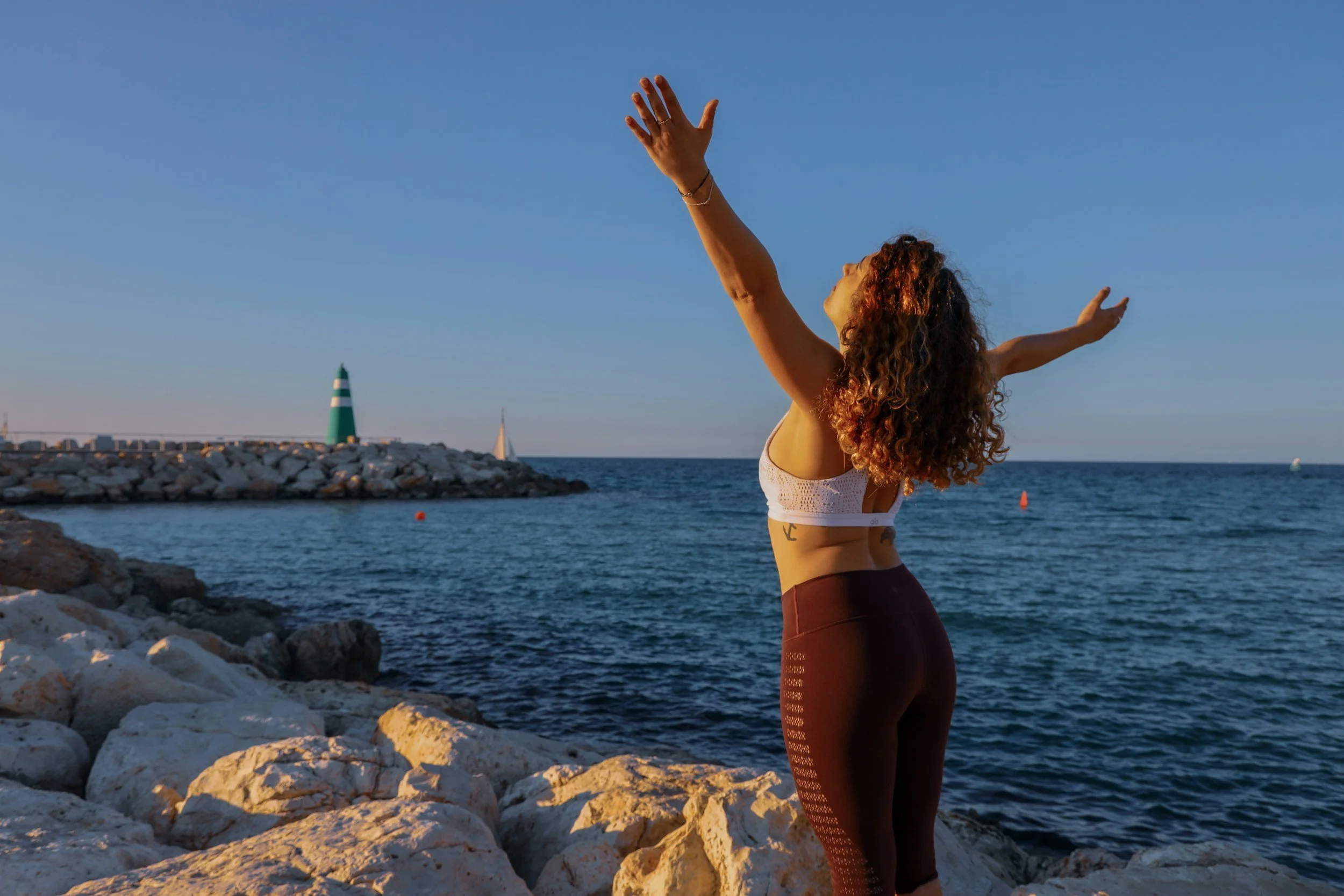 A woman with curly hair and tattoos on her side practicing yoga outdoors on rocks by the water during sunset, with her arms raised and palms open, facing the sky.