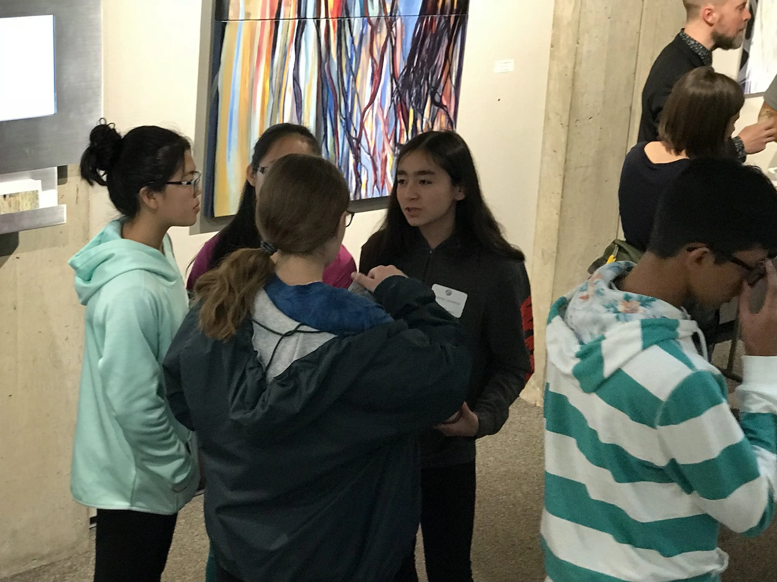 Group of young people talking in an art gallery with colorful abstract painting on the wall.