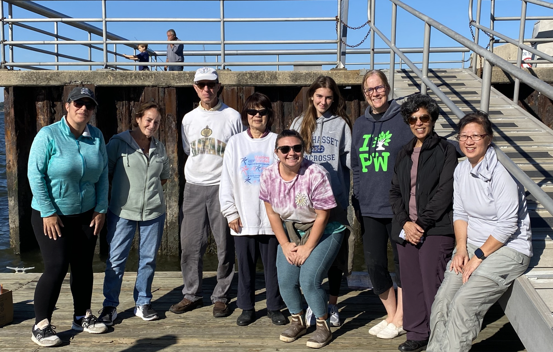 Group of people standing on a wooden dock near water, posing for a photo, some wearing sunglasses and casual outdoor clothing, with a railing and stairs behind them.